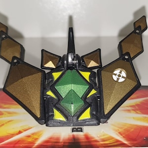 Pode incluir: Um brinquedo preto, verde e dourado com formas geométricas. O brinquedo tem uma forma central de diamante verde, rodeada por detalhes em preto e amarelo. Painéis dourados em forma de diamante se estendem para fora. Um pequeno emblema circular branco e dourado é visível.