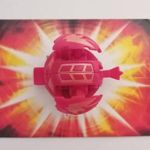 Op de afbeelding: Een roze en gouden Bakugan-speelgoed met een stervormige achtergrond. Het speelgoed is open en de kern is zichtbaar.