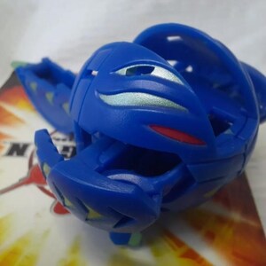 May include: A blue toy figure with a segmented design, featuring silver and red accents. The toy is partially open, revealing its internal structure. It rests on a colorful card with the letter "N" visible.