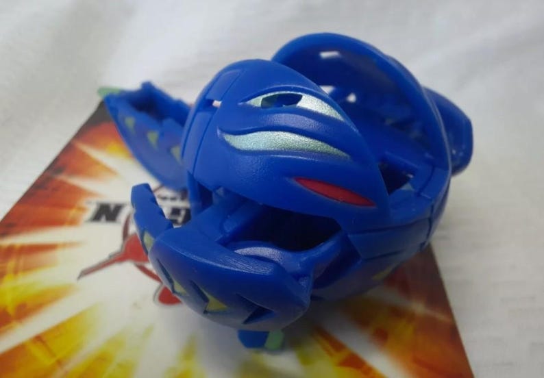 May include: A blue toy figure with a segmented design, featuring silver and red accents. The toy is partially open, revealing its internal structure. It rests on a colorful card with the letter "N" visible.