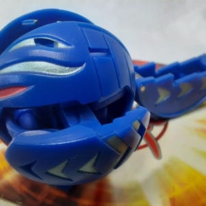 May include: A blue spherical toy with silver and red accents, partially open to reveal an interior. A detached blue piece with silver arrow-shaped designs is also present. The toy is on a colorful background.