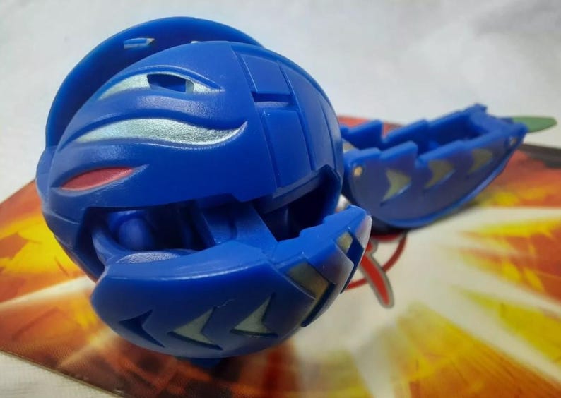 May include: A blue spherical toy with silver and red accents, partially open to reveal an interior. A detached blue piece with silver arrow-shaped designs is also present. The toy is on a colorful background.