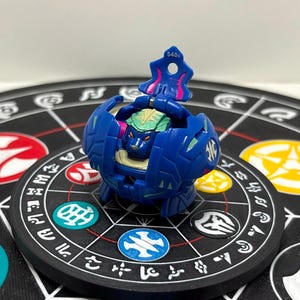 May include: A blue spherical toy with a mechanical design that opens to reveal a green and black creature head inside. The top section features a small hole and the printed text 540g. The toy has intricate moulded details and small pink accents.