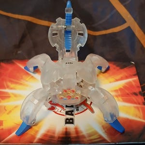 Puede incluir: Un juguete de plástico transparente con un diseño azul y plateado. El juguete tiene forma de escorpión y tiene el número "440" impreso en él. El juguete está sobre un fondo rojo y blanco con la palabra "Bakugan" impresa en él.