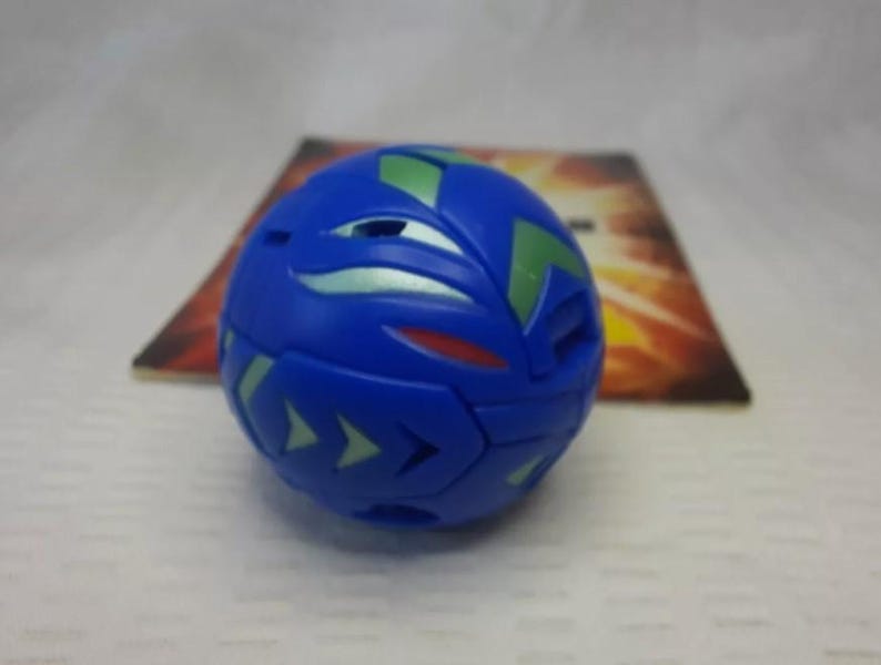 May include: A blue spherical toy with green and silver accents. The toy has a geometric design with arrow-shaped details. A colorful card is in the background.