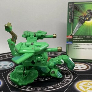 Op de afbeelding: Een groene speelgoedrobot met een gedetailleerd ontwerp, met twee kanonnen en beweegbare ledematen. Het speelgoed staat op een patroon, met een groene en witte kaart op de achtergrond. De robot heeft gouden accenten.