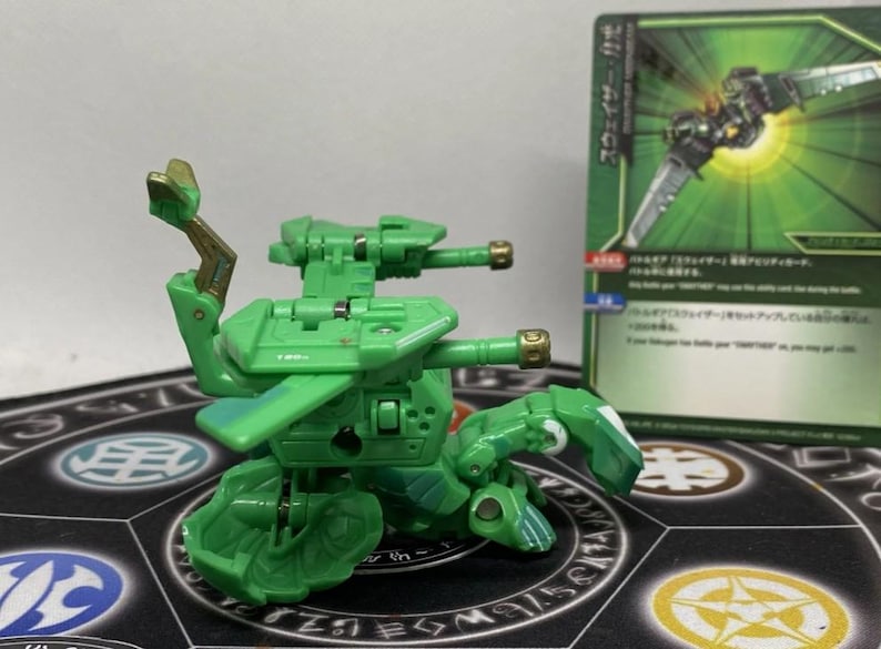 Op de afbeelding: Een groene speelgoedrobot met een gedetailleerd ontwerp, met twee kanonnen en beweegbare ledematen. Het speelgoed staat op een patroon, met een groene en witte kaart op de achtergrond. De robot heeft gouden accenten.