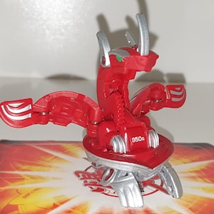 May include: Red and silver Bakugan toy with wings and a dragon-like head. The toy is posed on a red and yellow Bakugan card with the text "950g".