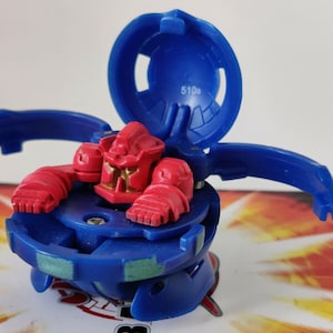 May include: A blue and red Bakugan toy with a red robot inside. The toy has a blue dome that opens to reveal the robot. The dome has the number 510b printed on it.