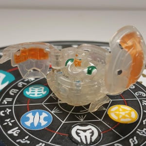 May include: A translucent toy figure with orange and green accents, partially open, resting on a black and white patterned surface with colorful circular tokens. The toy has a spherical shape with articulated wings.