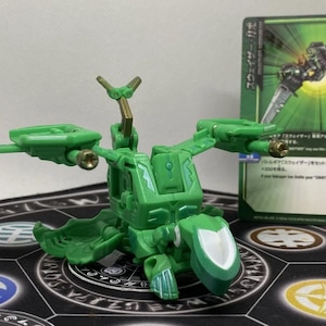 Op de afbeelding: Een groene speelgoedrobot met beweegbare vleugels en een wit schildachtig element. Het speelgoed staat voor een ruilkaart met Japanse tekst en een groen-zwarte afbeelding.