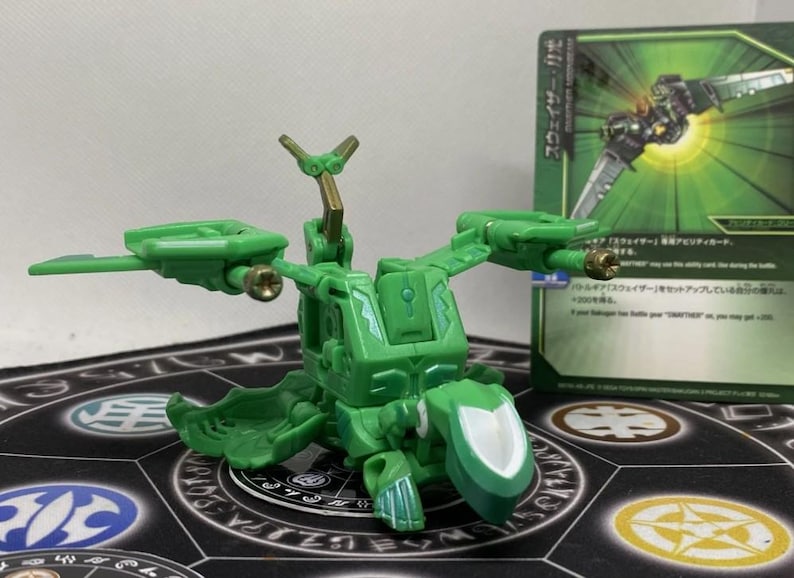 Op de afbeelding: Een groene speelgoedrobot met beweegbare vleugels en een wit schildachtig element. Het speelgoed staat voor een ruilkaart met Japanse tekst en een groen-zwarte afbeelding.