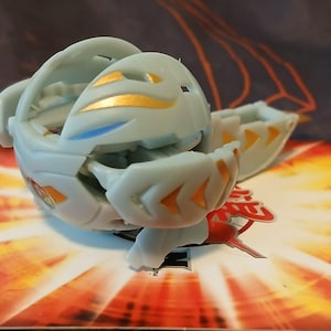 Puede incluir: Un Beyblade blanco y dorado con detalles azules. El Beyblade está en una posición parcialmente abierta, revelando el funcionamiento interno del juguete.