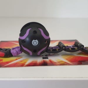 以下が含まれることがあります： 前面に白いシンボルが付いた黒と紫のBakuganのおもちゃ。おもちゃは閉じたボールの形をしていて、黒と金の延長アームが付いています。