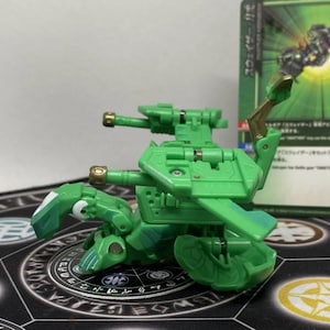 Op de afbeelding: Een groene speelgoedrobot met twee kanonnen en een vleugelachtige structuur. Het speelgoed staat op een patroonmat, met een ruilkaart op de achtergrond. De robot heeft goudkleurige accenten.