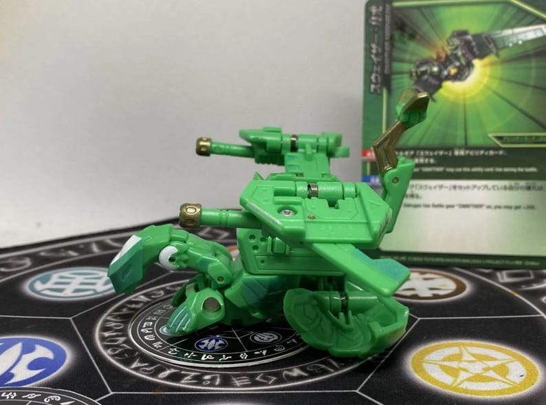 Op de afbeelding: Een groene speelgoedrobot met twee kanonnen en een vleugelachtige structuur. Het speelgoed staat op een patroonmat, met een ruilkaart op de achtergrond. De robot heeft goudkleurige accenten.