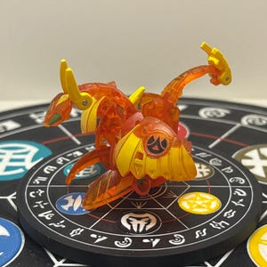 Puede incluir: Una figura de dragón de juguete translúcida naranja y amarilla con extremidades articuladas. El juguete está posado sobre una plataforma circular negra con símbolos y patrones coloridos. El dragón tiene un diseño detallado con bordes afilados y una pose dinámica.