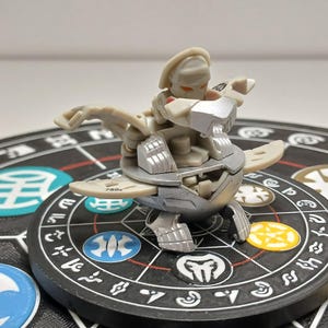 Puede incluir: Una figura de juguete Bakugan gris claro con detalles plateados, colocada sobre un tablero de juego negro y gris. La figura tiene un diseño detallado con partes articuladas y está en una pose dinámica. El tablero de juego presenta diseños circulares coloridos.