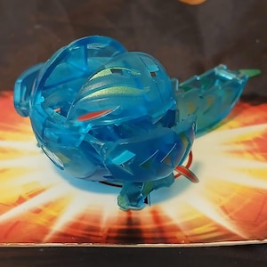 Puede incluir: Un juguete azul translúcido con forma esférica, con detalles rojos y verdes. El juguete está parcialmente abierto, revelando una estructura interna. Descansa sobre una superficie colorida y estampada con tonos naranja y amarillo.