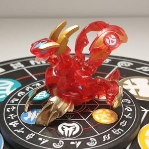 以下が含まれることがあります： 頭部、尾、脚に金色のアクセントが施された、赤く半透明なドラゴン型の玩具フィギュア。このフィギュアは関節部分が可動し、尾には円形のエンブレムが付いています。白いシンボルが描かれた黒い円形の台座の上に置かれています。