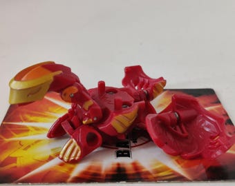 May include: Red and gold plastic toy with a scorpion design. The toy has a segmented body and tail, with a large head and pincers. The toy is on a red and yellow background with the word "Bakugan" printed on it.