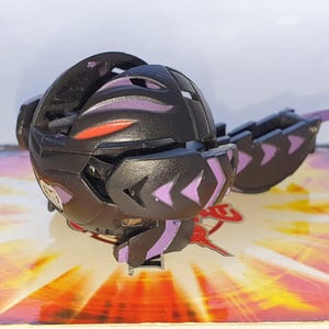 Puede incluir: Una figura de juguete negra con detalles morados y rojos, con un diseño segmentado. El juguete está parcialmente abierto, revelando detalles internos. Descansa sobre una superficie colorida con un logotipo.