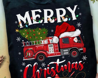 May include: Black t-shirt with a Christmas-themed design. The graphic shows a red fire engine with a Christmas tree, Santa Claus, and the words "Merry Christmas" in white and red. Snowflakes and fairy lights add to the festive feel.