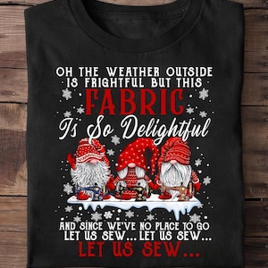 May include: Black t-shirt with the text "OH THE WEATHER OUTSIDE IS FRIGHTFUL BUT THIS FABRIC Is So Delightful." Below the text are three gnomes wearing red hats and holding sewing machines. The shirt is folded.