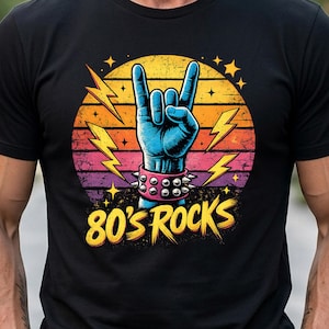 May include: A black t-shirt featuring a graphic of a blue hand making a rock-and-roll gesture, wearing a pink studded wristband. The design includes yellow lightning bolts, a sunset-colored circle background, and the text 80'S ROCKS in yellow.