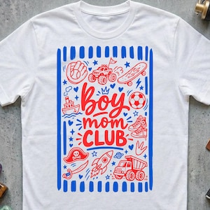 May include: A white short-sleeved t-shirt featuring a central rectangular graphic with blue vertical stripes on the sides. The red text reads Boy mom CLUB, surrounded by illustrations of a baseball glove, monster truck, skateboard, boat, football, trainers, pirate hat, sword, rocket, and dump truck.