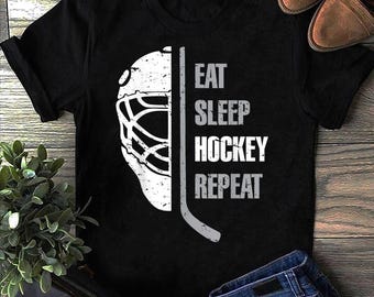 May include: Black t-shirt featuring a white hockey goalie mask graphic and a hockey stick. The text "EAT SLEEP HOCKEY REPEAT" is printed vertically on the right side of the shirt. The shirt is made of a soft material.