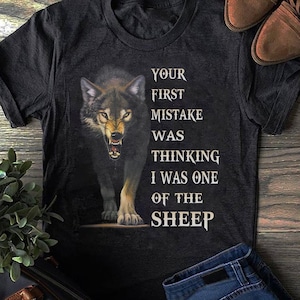 Puede incluir: Camiseta gris oscuro con un lobo gruñendo. El texto dice: "YOUR FIRST MISTAKE WAS THINKING I WAS ONE OF THE SHEEP." La camiseta tiene mangas cortas y cuello redondo.