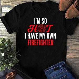 May include: A black short-sleeved t-shirt featuring white and red text that reads I'M SO HOT I HAVE MY OWN FIREFIGHTER, with a red firefighter department emblem replacing the letter O in the word HOT, printed on the front of the garment.