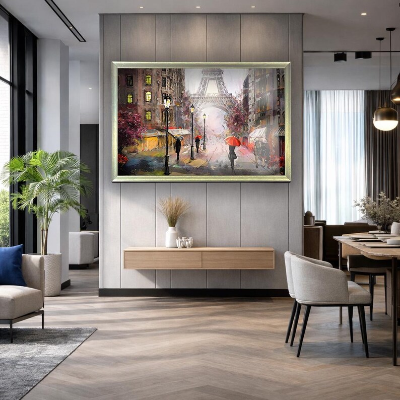 May include: A framed painting of a Parisian street scene with the Eiffel Tower in the background. The artwork depicts figures with umbrellas walking along a wet street, with buildings and streetlights lining the road. The painting is displayed in a modern living room.