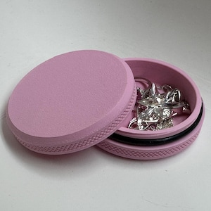 May include: A light pink, round jewelry box with a textured lid and a black ring around the base. The box is open, revealing several silver rings and necklaces inside. The box is on a white surface.