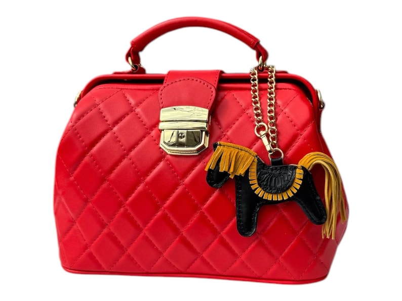 Puede incluir: Un bolso de mano de estilo bolso de m&eacute;dico acolchado rojo con cierre dorado y un dije de caballo negro y dorado sujeto a una cadena dorada.