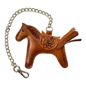 May include: A brown leather horse keyring with a gold chain. The horse has a saddle with a floral design and a fringe tail.