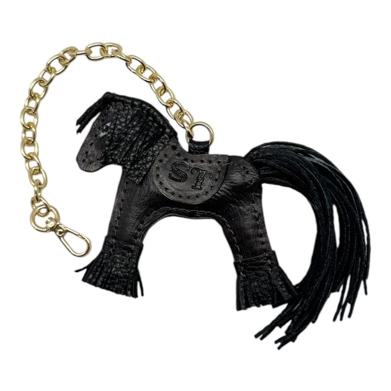 Op de afbeelding: Zwarte leren paarden sleutelhanger met een goudkleurige ketting en sluiting. Het paard heeft een getextureerde uitstraling, een lange zwarte manen en staart, en een zadel met de initialen "ST".