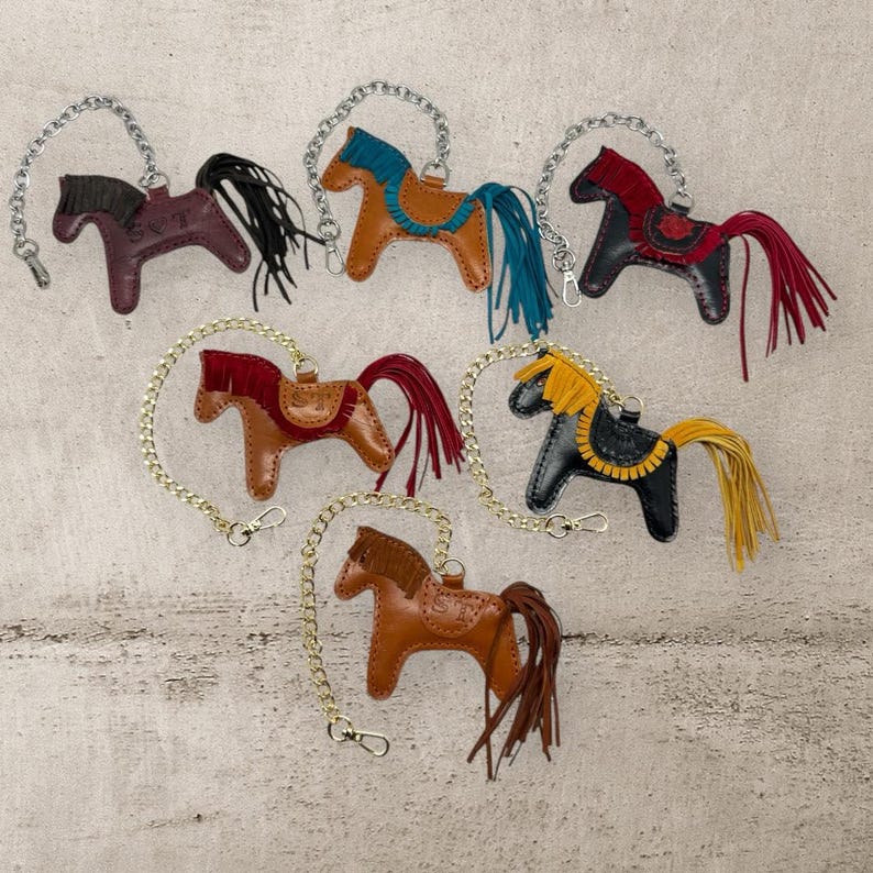 Puede incluir: Seis llaveros de cuero con forma de caballo con detalles de flecos. Cada llavero presenta un caballo de color diferente con una cadena y un cierre de metal. Los caballos son marrones, negros, rojos, tostados y azules.