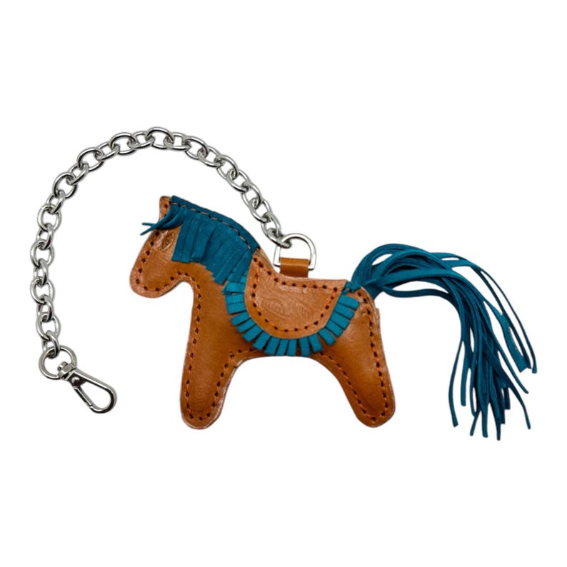 Puede incluir: Un llavero de cuero marr&oacute;n en forma de caballo con una crin y una cola de flecos azul turquesa. El llavero tiene una cadena plateada.