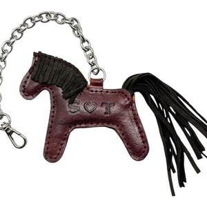 Puede incluir: Un llavero de cuero burdeos en forma de caballo con una cola de flecos negros y una cadena plateada. El caballo tiene una crin de gamuza negra y las letras "SOT" est&aacute;n grabadas en el lateral con un s&iacute;mbolo de coraz&oacute;n.
