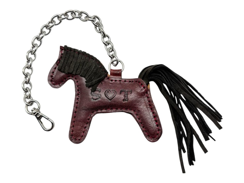 Puede incluir: Un llavero de cuero burdeos en forma de caballo con una cola de flecos negros y una cadena plateada. El caballo tiene una crin de gamuza negra y las letras "SOT" est&aacute;n grabadas en el lateral con un s&iacute;mbolo de coraz&oacute;n.