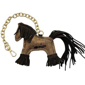 Op de afbeelding: Een bruin en goud paardvormige sleutelhanger met een gouden ketting en sluiting. Het paard heeft zwarte franjes voor de manen, staart en benen. Het lichaam van het paard heeft een distressed gouden metallic afwerking.