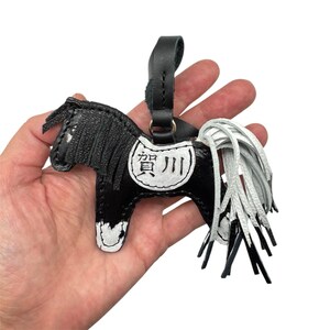 Op de afbeelding: Zwart en witte leren paarden sleutelhanger met een zwarte riem. Het paard heeft een wit zadel met Japanse karakters. De manen en staart zijn wit. De sleutelhanger wordt in een hand gehouden.