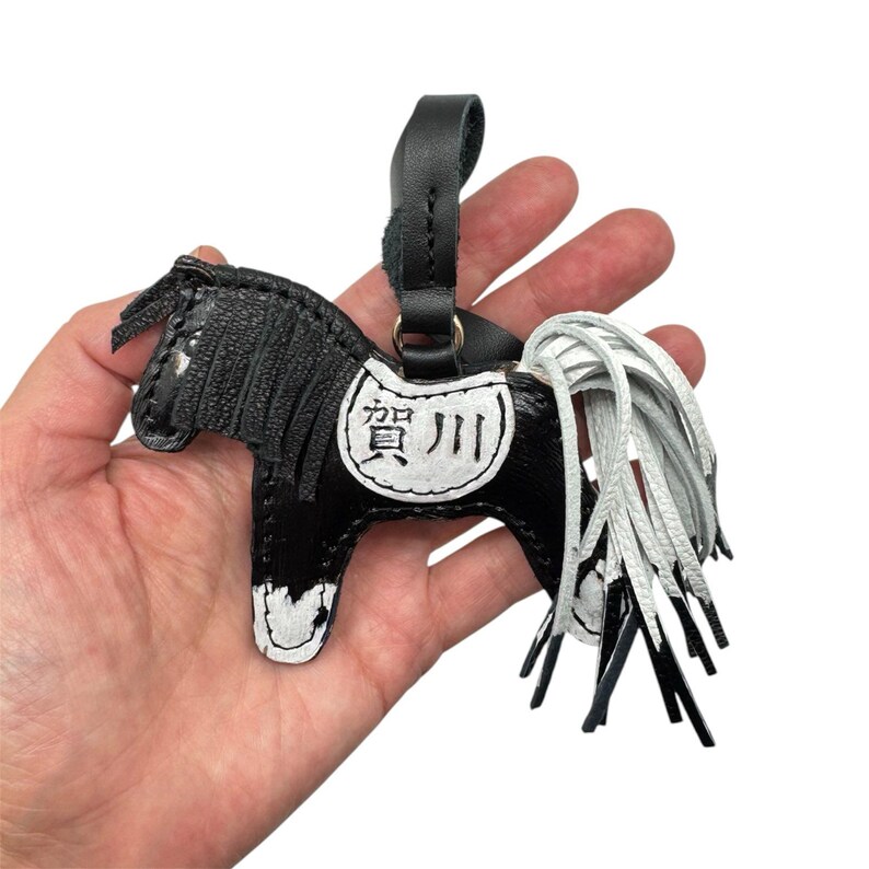 Op de afbeelding: Zwart en witte leren paarden sleutelhanger met een zwarte riem. Het paard heeft een wit zadel met Japanse karakters. De manen en staart zijn wit. De sleutelhanger wordt in een hand gehouden.