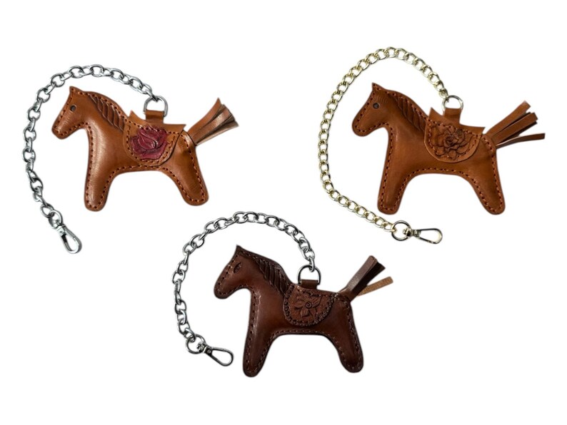 May include: Three brown leather horse keychains with silver and gold chains. Each horse has a saddle with a floral design.