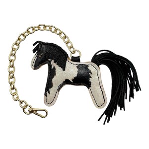 Op de afbeelding: Een leren paarden sleutelhanger met een gouden ketting en sluiting. Het paard heeft een zwart-wit vlekkenpatroon, een zwarte manen en een zwarte kwaststaart. Een decoratief accessoire.