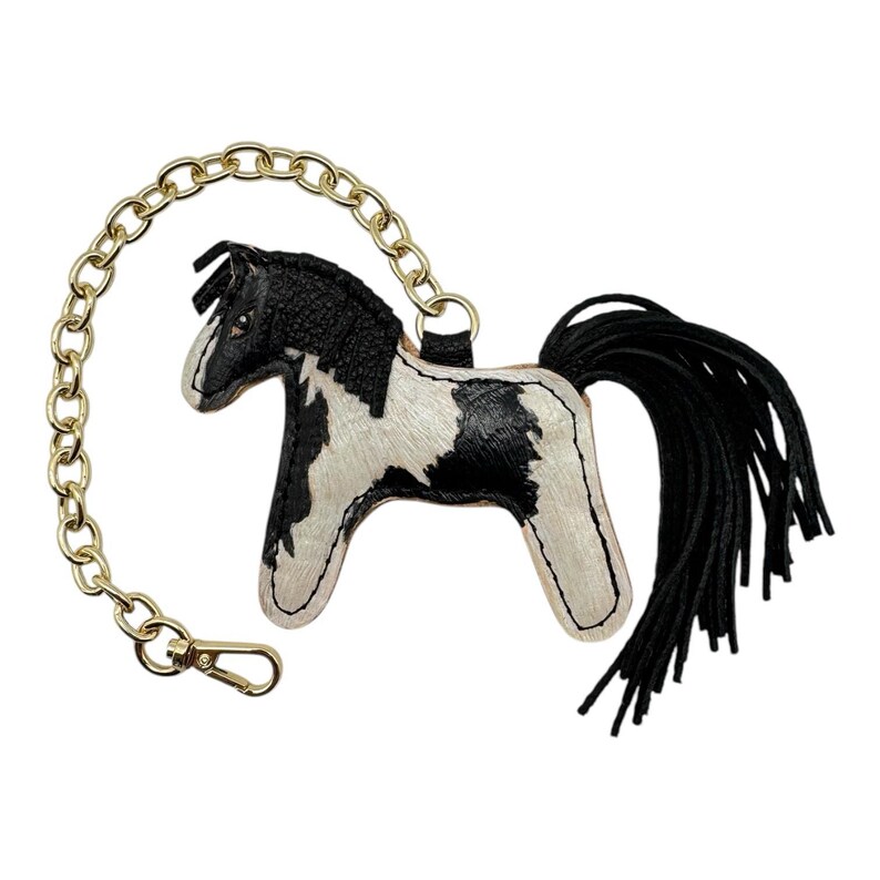 Op de afbeelding: Een leren paarden sleutelhanger met een gouden ketting en sluiting. Het paard heeft een zwart-wit vlekkenpatroon, een zwarte manen en een zwarte kwaststaart. Een decoratief accessoire.