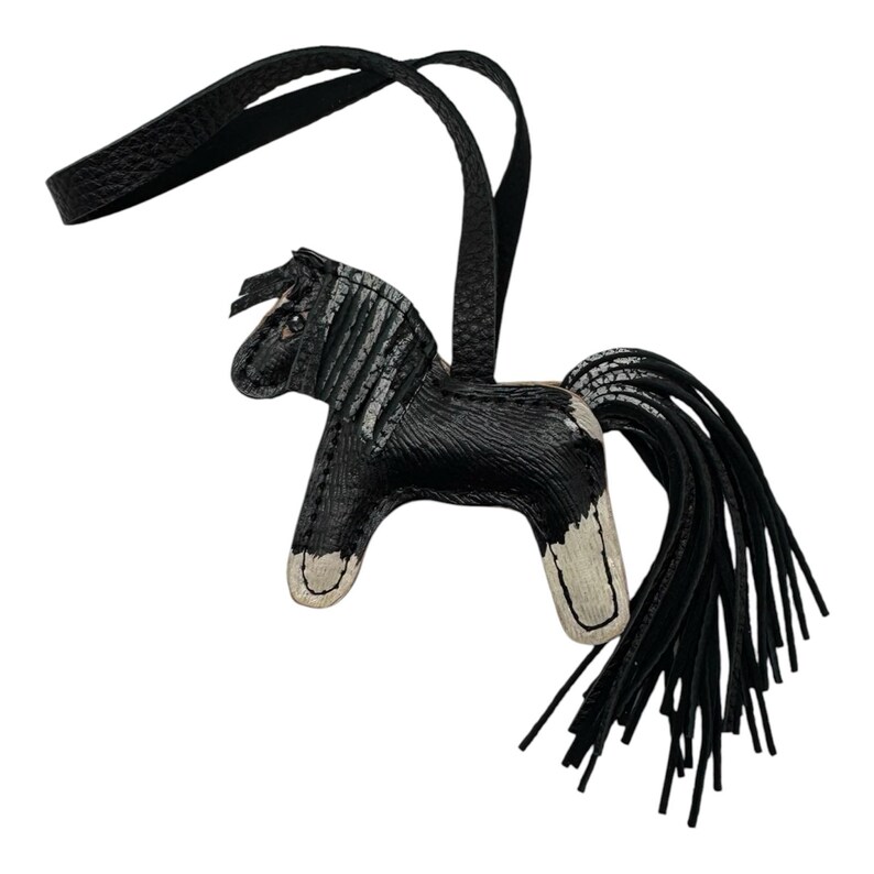 Op de afbeelding: Een zwart-witte leren paardvormige bedel met een lange zwarte staart en een zwarte riem. Het paard heeft witte accenten op de poten en een gestreepte manen. De bedel is waarschijnlijk een decoratief accessoire.