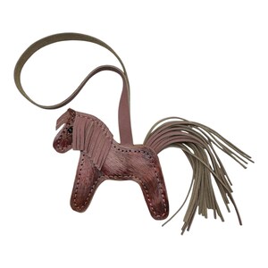 Op de afbeelding: Een leren paardenhanger in bruine en mauve tinten. Het paard heeft een gefranjerde manen en een lange, vloeiende staart. Er is een leren lus bevestigd om op te hangen. Deze hanger is waarschijnlijk een decoratief accessoire.